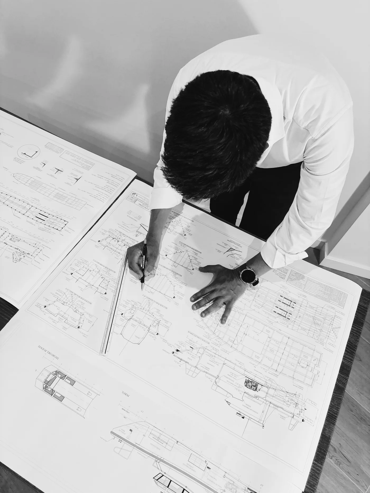Ingeniero naval trazando líneas sobre los planos de una embarcación, vista cenital en blanco y negro.